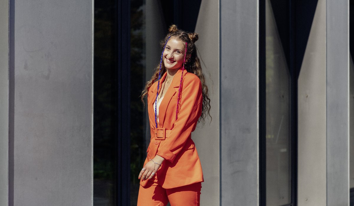 Mahalia ist Ausbildungsbotschafterin. Der IHK Azubi Kampagne könnenlernen. Sie hat lange braune Locken. Sie trägt einen Blazer und eine Hose in der Farbe Orange.