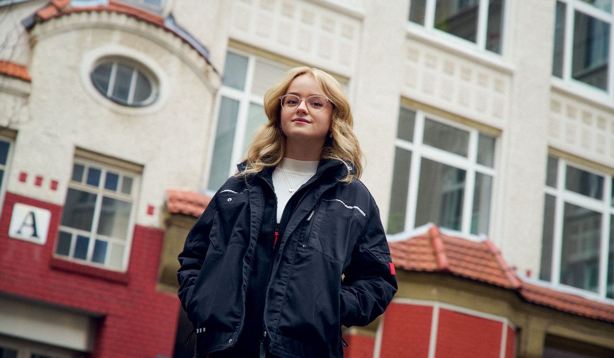 Emily ist Botschafterin für die Ausbildung. Sie hat blonde Haare und trägt eine Brille. Emeily steht draßen vor einem Haus und trägt eine dunkle Jacke.