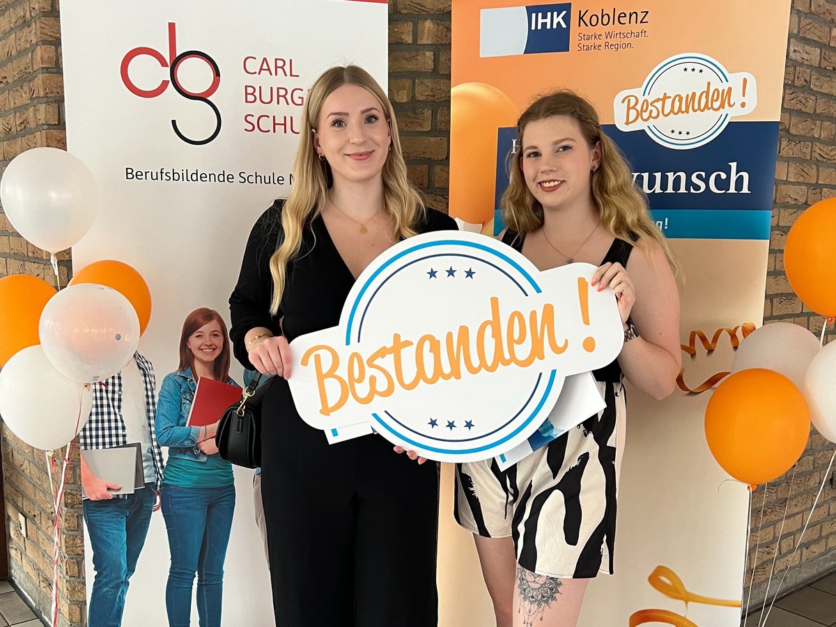 Azubi Paula und Lea stehen vor einem Rollup und halten ein Schild mit der Aufschrift Bestanden hoch. Sie haben ihre Ausbildung erfolgreich abgeschlossen.