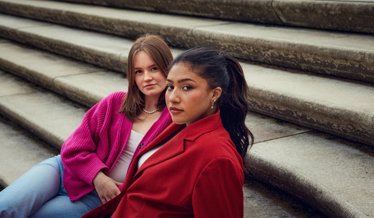 Selina und Anna sind Botschafterinnen für die Ausbildung. Beide sitzen auf einer Treppe. Selina trägt einen roten Blazer und hat schwarze Haare. Haare trägt eine pinke Jacke und hat braune Haare.