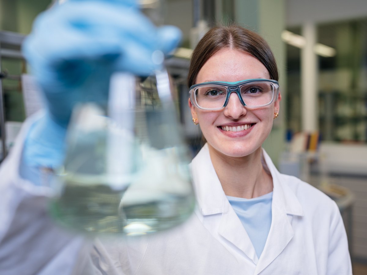 Taminas Ausbildungsberuf ist Chemielaborantin. Sie steht in einem Labor und trägt eine Schutzbrille und einen Kittel. In der Hand hält sie ein Reagenzglas..
