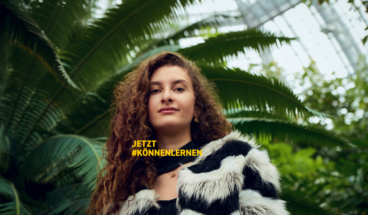 Mahalia ist Ausbildungsbotschafterin. Der IHK Azubi Kampagne könnenlernen. Sie hat lange braune locken und steht vor einer großen Palme. .