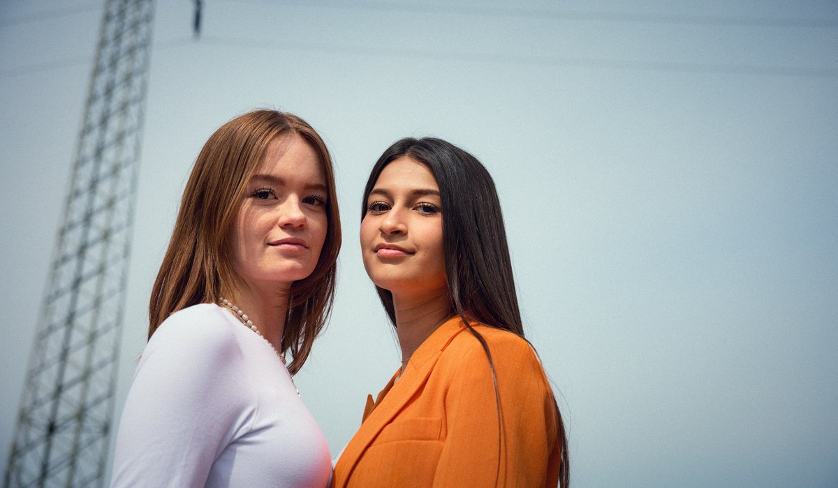 Aylin und Anna sind Botschafter für die Ausbildung. Beide stehen draußen an einem Strommaßt. Anna trägt ein weißes Oberteil und Aylin eine Jacke in orange.