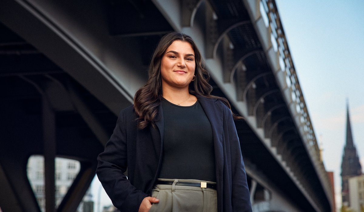 Feyza ist Botschafterin für die Ausbildung. Sie steht draußen vor einer dunklen Brücke. Feyza hat schwarze Haare und trägt ein schwarzes Oberteil und schwarzen Blazer. Sie posiert draußen vor der Brücke. 