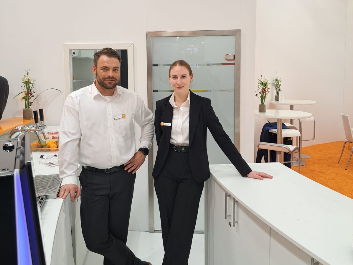 Azubi Megan und Azubi Timo haben sich schick für eine Eisenbahnmesse angezogen. Timo trägt eine schwarze Hose und ein weißes Hemd. Megan trägt eine weiße Bluse und eine schwarze Hose sowie eine schwarze Bluse.